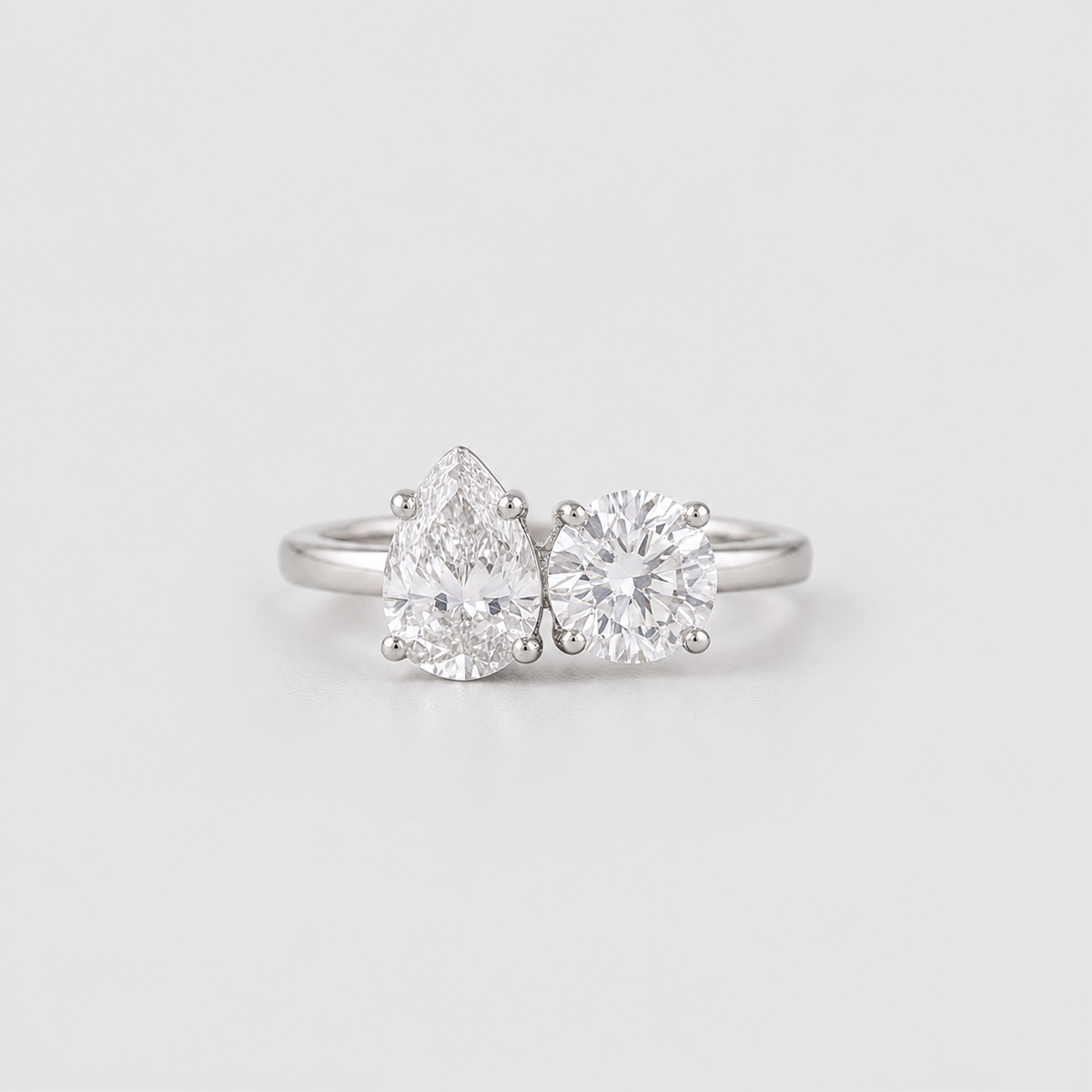 A silver engagement ring featuring a pear-cut diamond on the left and a round-cut diamond on the right, set on a polished silver band against a light gray background.