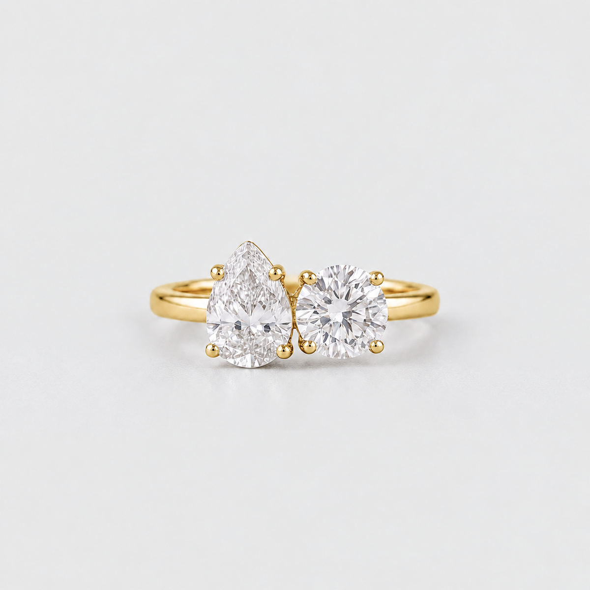 A gold ring featuring a pear-cut diamond on the left and a round-cut diamond on the right, set on a plain light gray background.