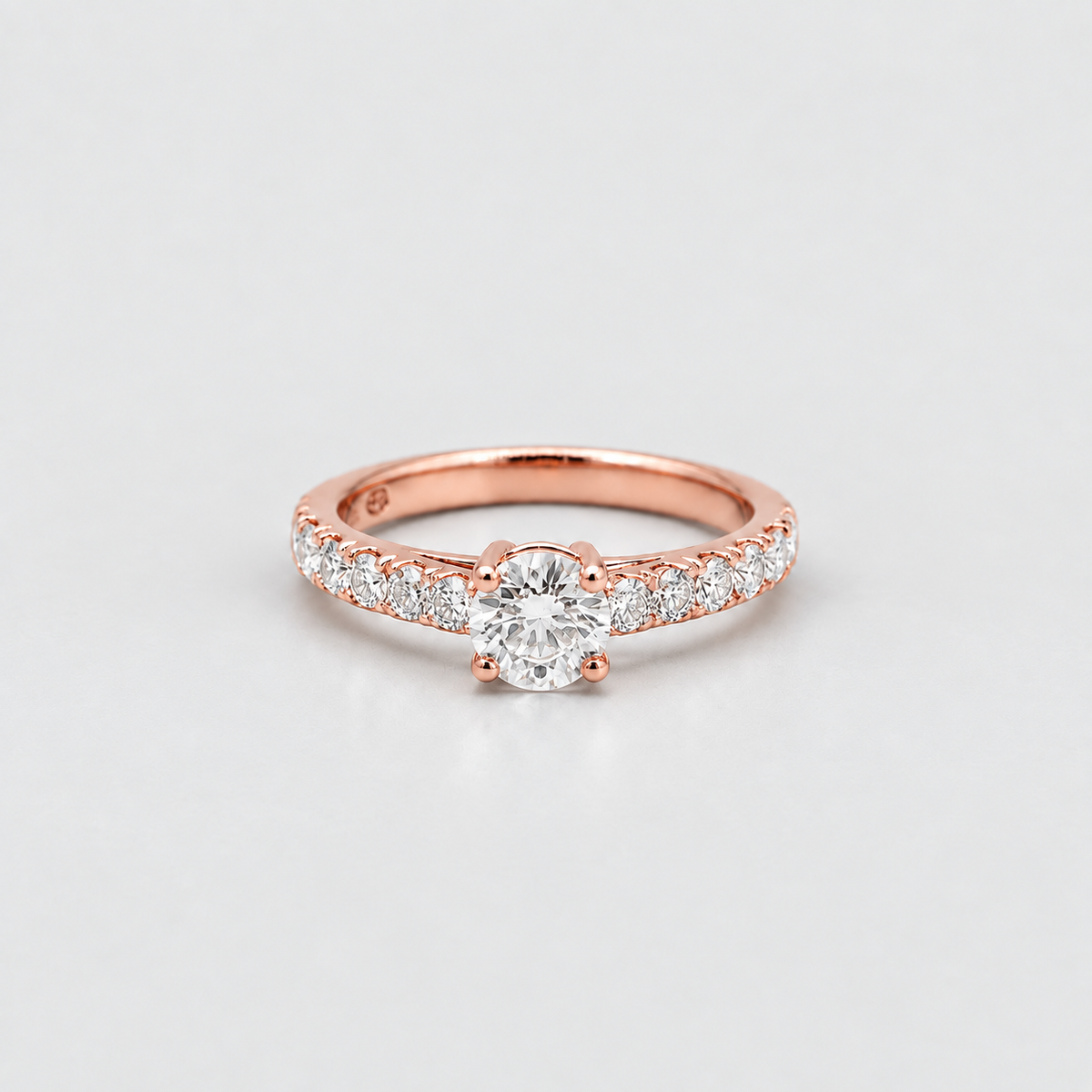 A rose gold solitaire engagement ring with a round-cut center stone and pavé-set diamonds along the shank, photographed on a clean light gray background.