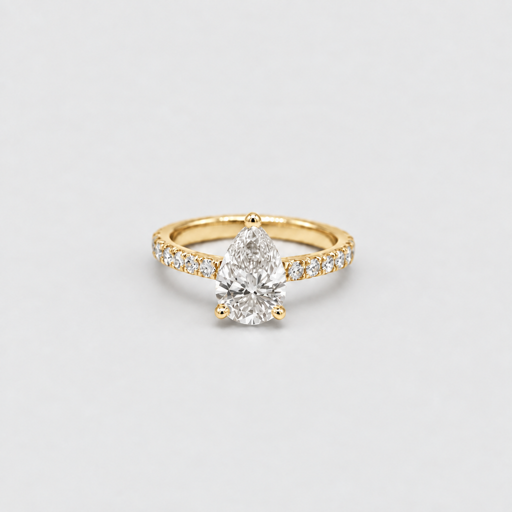 A yellow gold ring featuring a pear-cut diamond center stone and pavé-set diamonds along the band, photographed on a clean, light gray background.