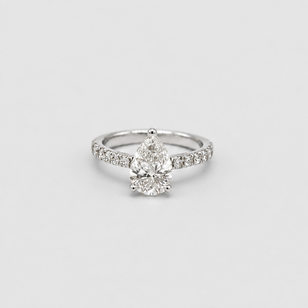 A silver-toned diamond ring featuring a pear-cut center stone and a pavé band with smaller diamonds, set against a plain light gray background.