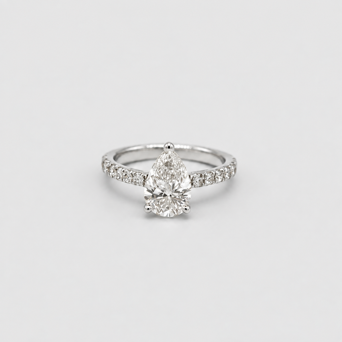 A silver-toned diamond ring featuring a pear-cut center stone and a pavé band with smaller diamonds, set against a plain light gray background.