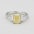 A yellow diamond engagement ring with a rectangular radiant-cut center stone and baguette-cut side stones on a polished silver-colored band, set against a light gray background.