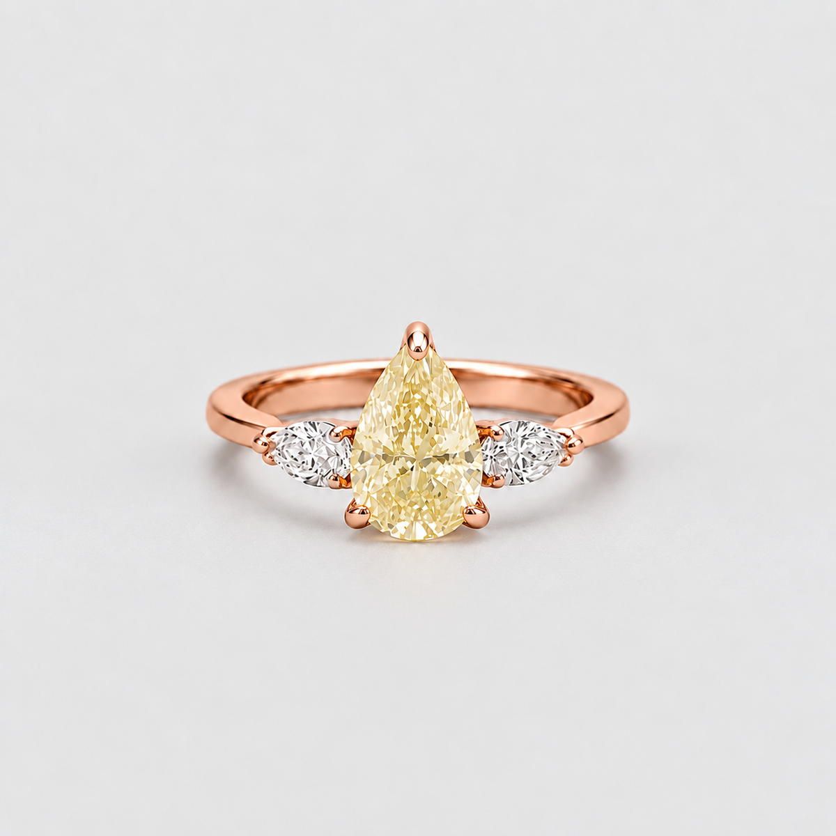 A rose gold ring featuring a pear-cut yellow center stone flanked by two small round clear stones, set against a clean light gray background.