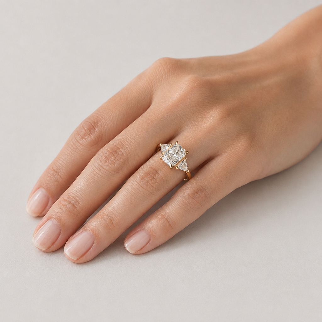 A close-up of a hand wearing a yellow gold ring with a pear-cut diamond center stone and smaller diamonds on the shoulders, set against a neutral light gray background.