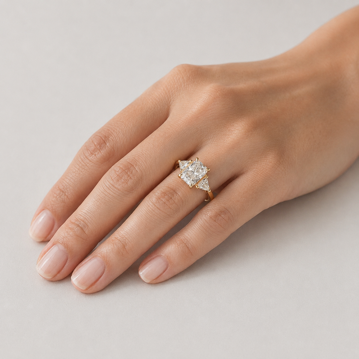 A close-up of a hand wearing a yellow gold ring with a pear-cut diamond center stone and smaller diamonds on the shoulders, set against a neutral light gray background.