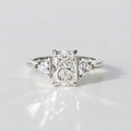 A three-stone diamond engagement ring with a central rectangular radiant-cut diamond flanked by two smaller round-cut diamonds on a polished silver-colored band, set against a bright white background with a soft reflection.