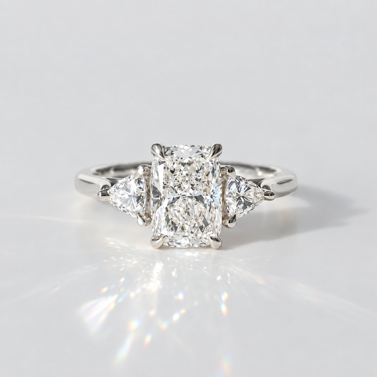 A three-stone diamond engagement ring with a central rectangular radiant-cut diamond flanked by two smaller round-cut diamonds on a polished silver-colored band, set against a bright white background with a soft reflection.