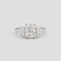 A three-stone diamond engagement ring with a central radiant-cut diamond flanked by two smaller cushion-cut diamonds on a polished silver-colored band, set against a light gray background.