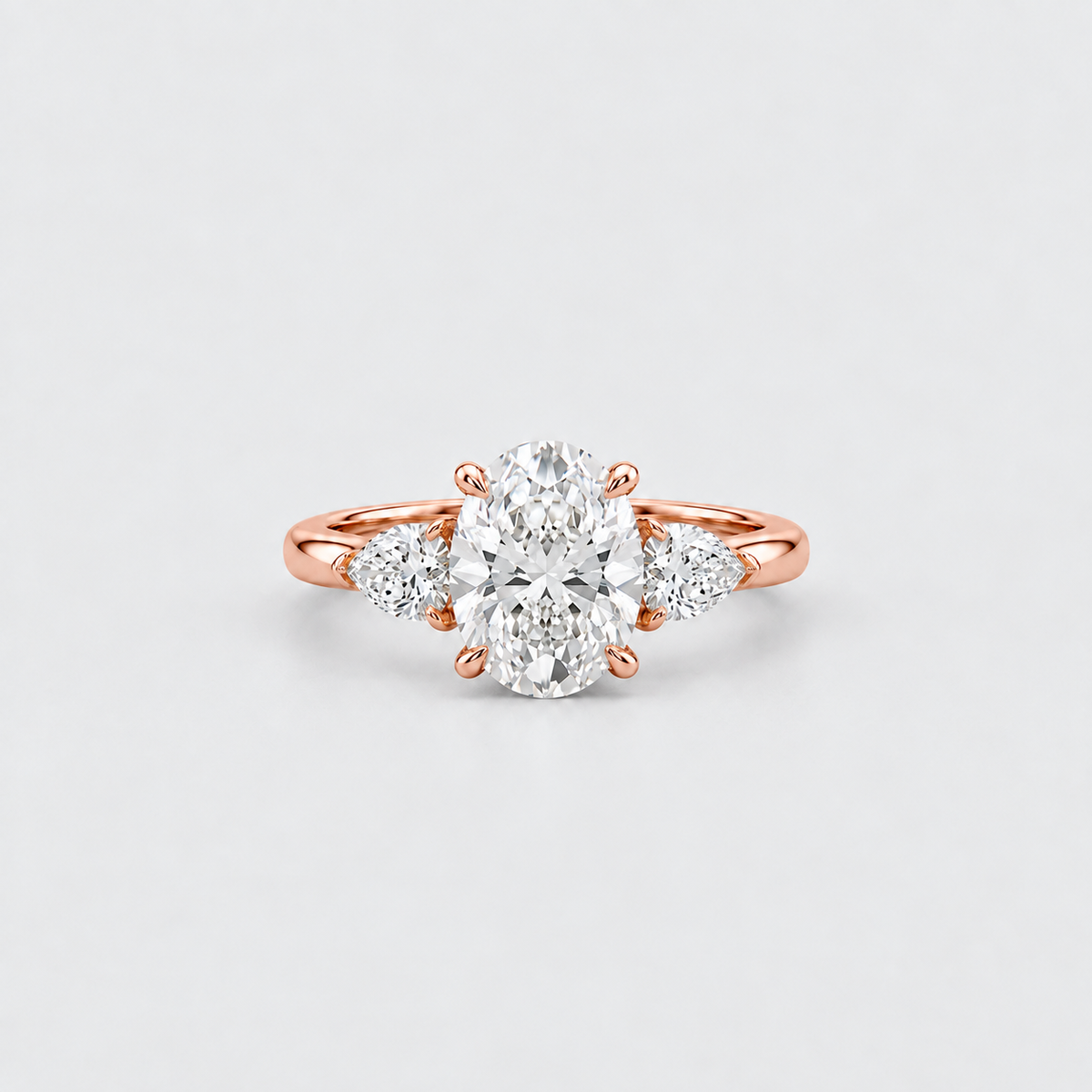 A rose gold engagement ring featuring a large oval center diamond flanked by two round side diamonds, set against a clean light gray background.