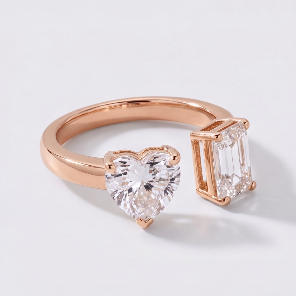 A rose gold ring featuring a heart-shaped clear gemstone and an emerald-cut clear gemstone, photographed on a clean white background.