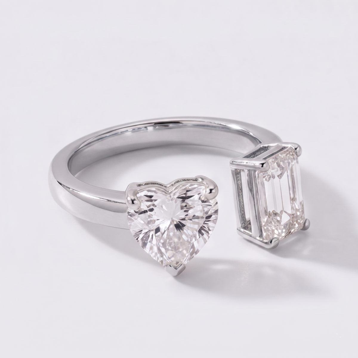 A silver-toned ring featuring a large heart-shaped clear gemstone on one side and an emerald-cut clear gemstone on the other, photographed on a clean white background.