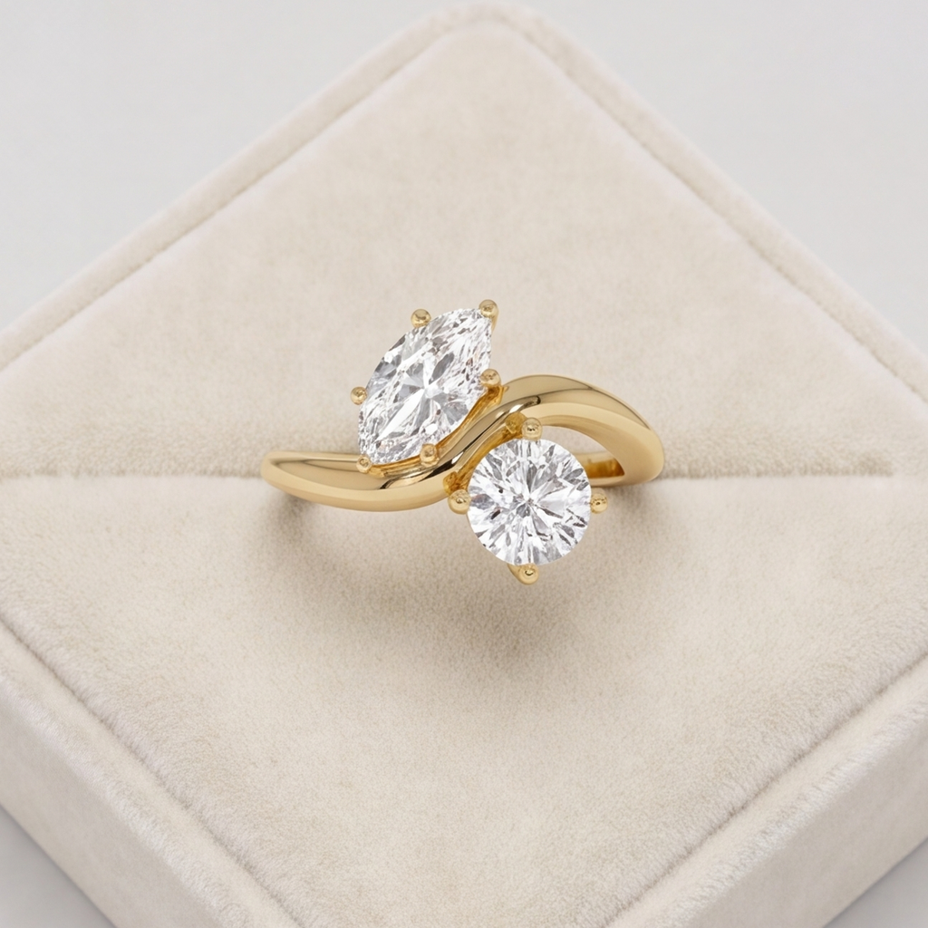 A yellow gold ring with two clear gemstones, one marquise-cut and one round-cut, displayed on a cream-colored velvet ring box.