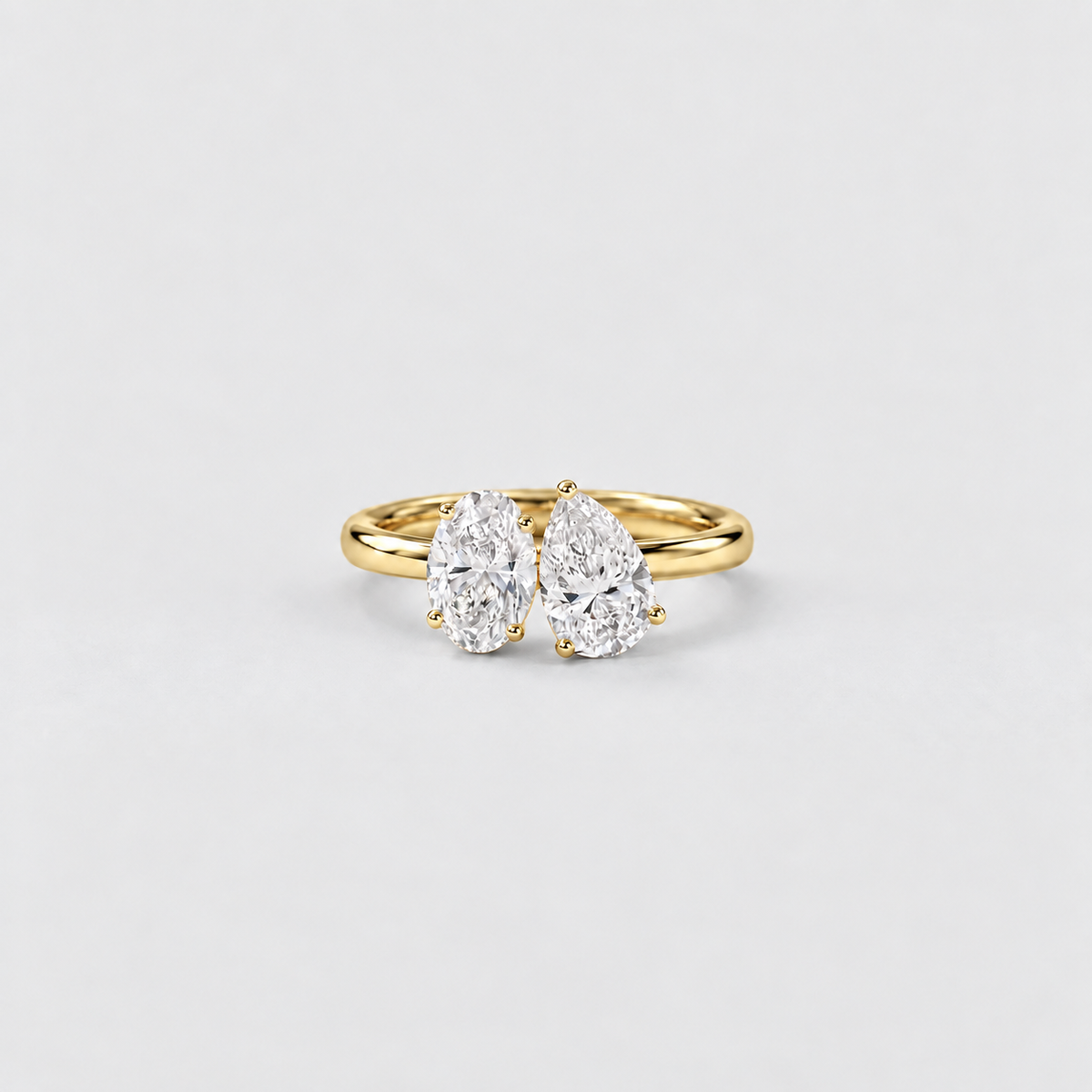 A yellow gold ring featuring two pear-cut diamonds in a side-by-side setting, photographed on a clean, light gray background.