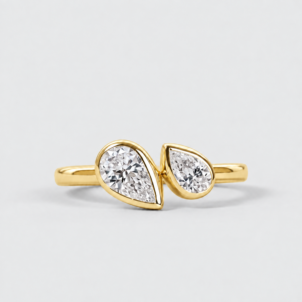 A gold ring featuring two pear-cut clear gemstones in bezel settings, photographed on a clean light gray background.