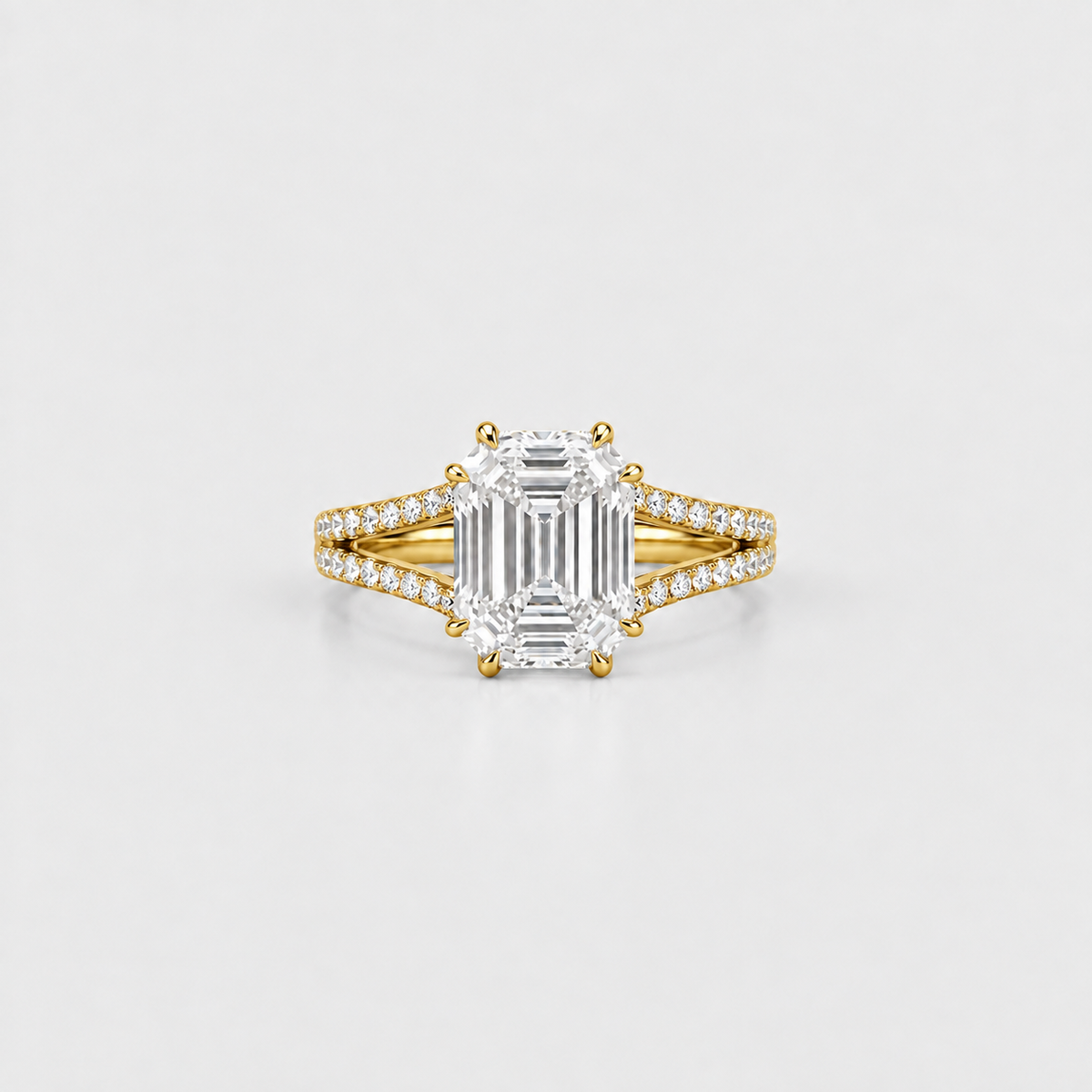 An emerald-cut diamond ring with a yellow gold band accented by pavé diamonds, centered on a light gray background.