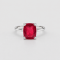 A silver ring featuring a cushion-cut, faceted red gemstone in a four-prong setting, photographed on a clean, light gray background.