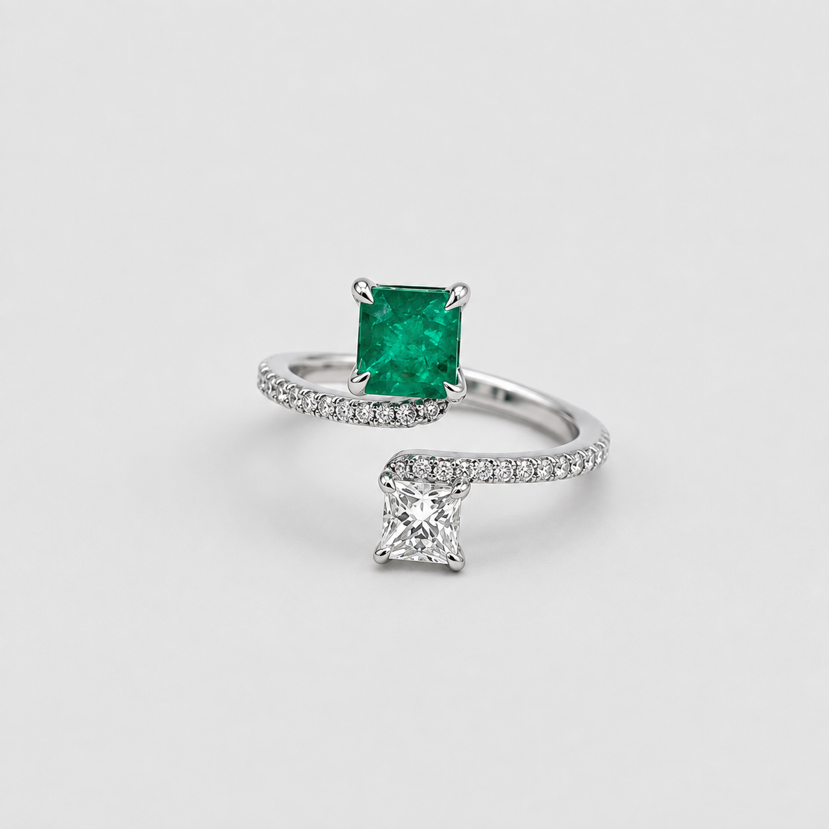 A silver-tone ring featuring a square-cut green emerald gemstone and a separate princess-cut clear gemstone, both set on a pavé-set silver band, photographed on a light gray background.