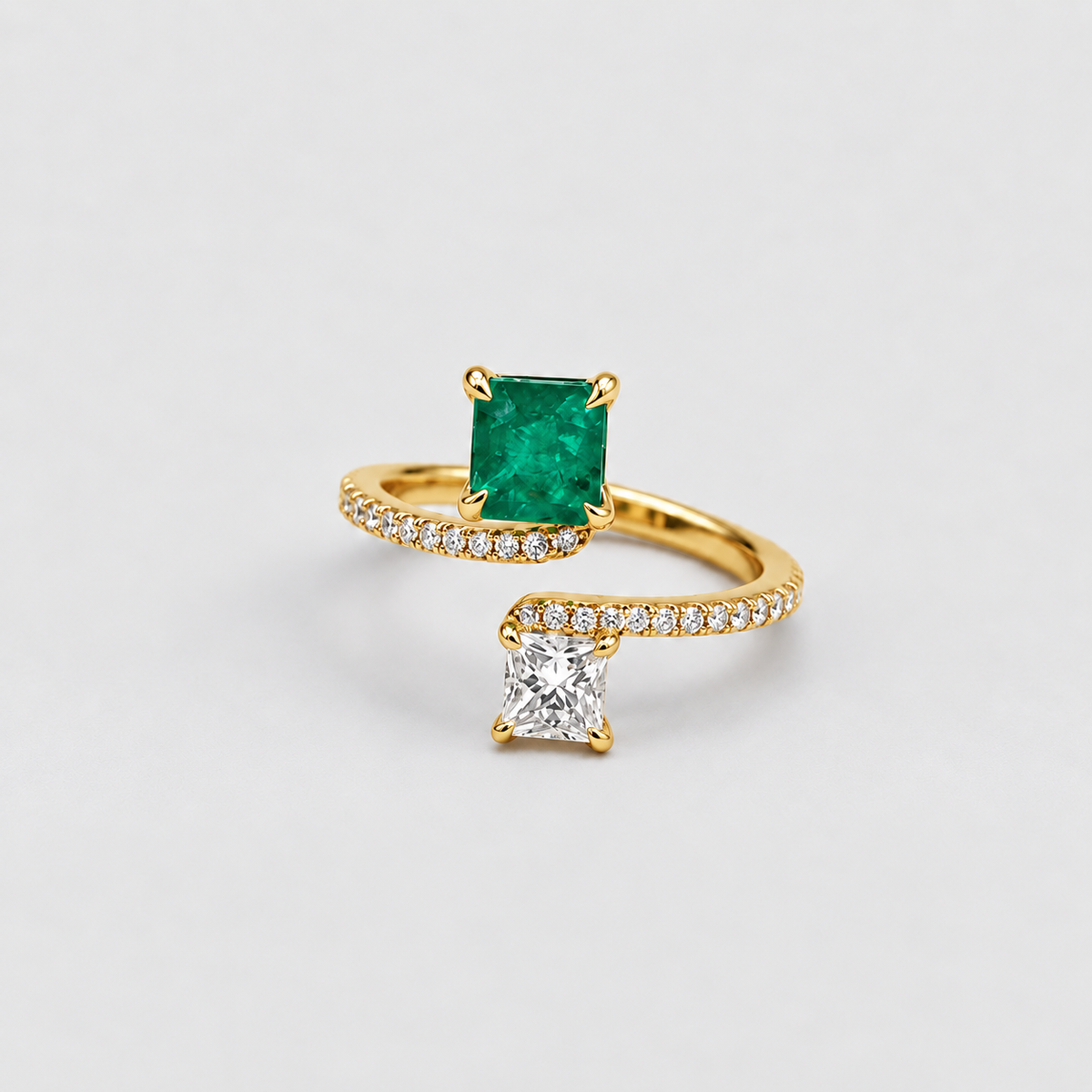 A gold ring featuring a square-cut green emerald gemstone and a separate square-cut clear diamond, both suspended from pavé-set gold bands, photographed on a light gray background.