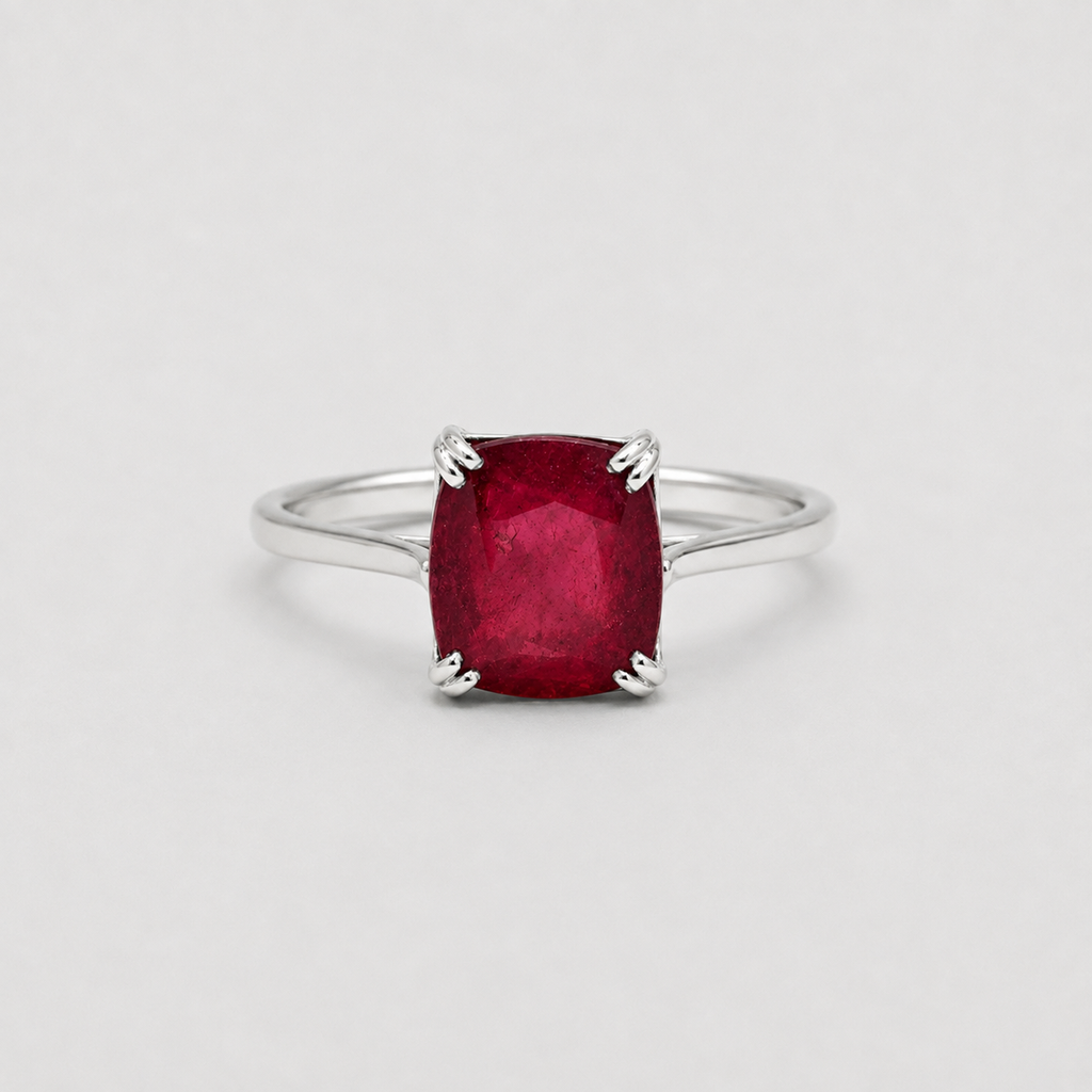 A silver ring featuring a cushion-cut, faceted deep red gemstone in a four-prong setting, shown against a neutral light gray background.