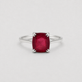 A silver ring featuring a cushion-cut, faceted deep red gemstone in a four-prong setting, shown against a neutral light gray background.