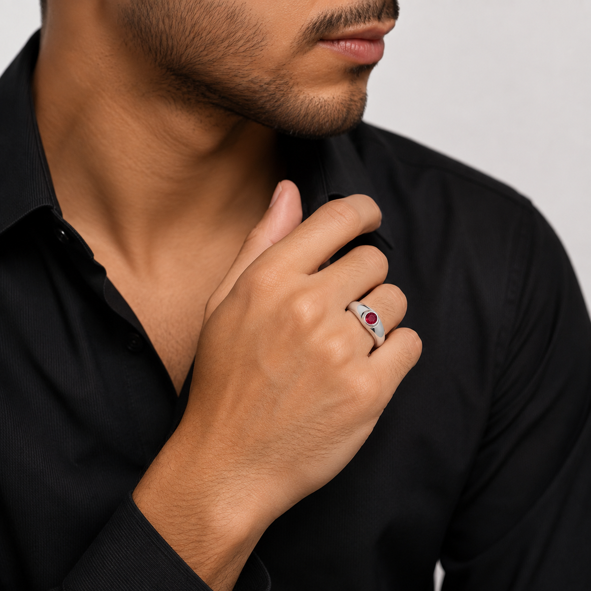 A close-up of a man wearing a silver-toned ring with a red oval gemstone on his ring finger, dressed in a black collared shirt against a light gray background.