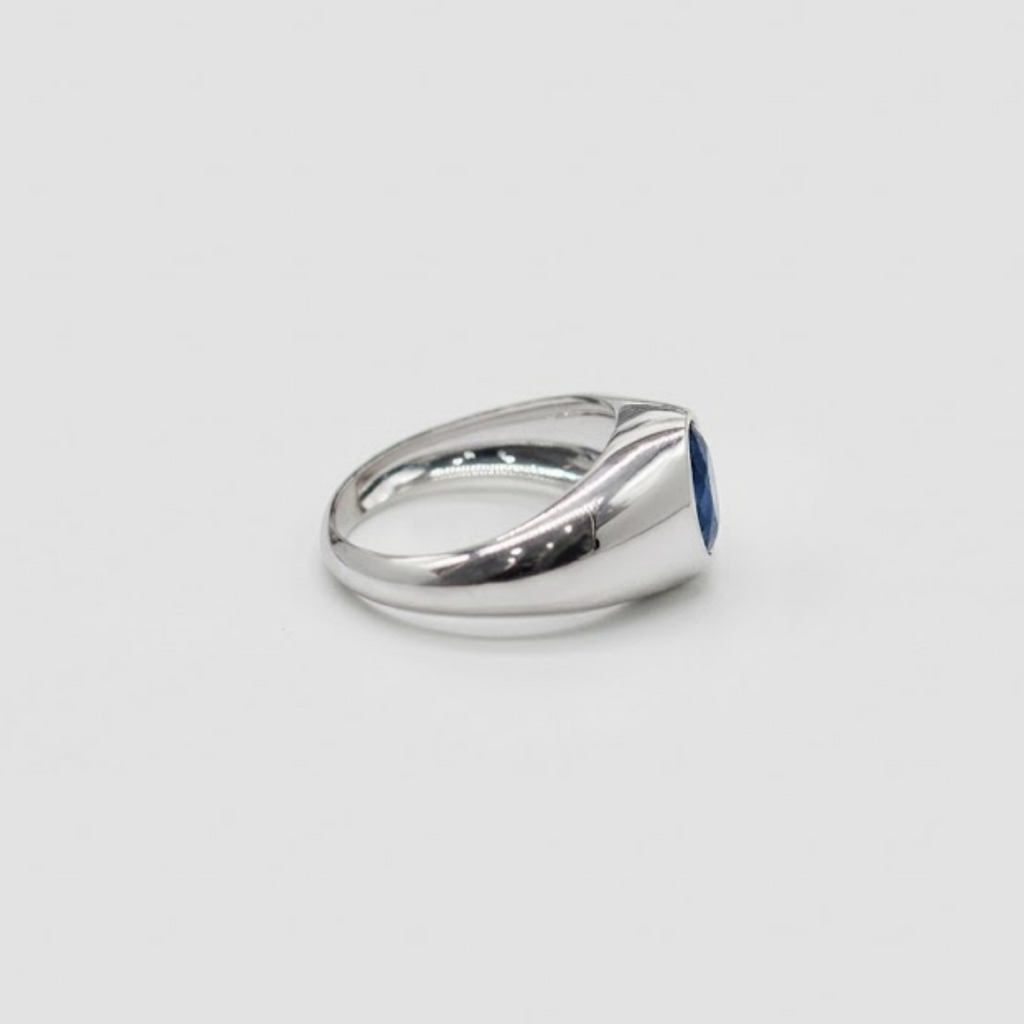A polished silver ring featuring a small, round blue gemstone, photographed on a neutral light gray background.