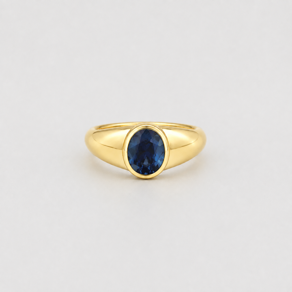 A yellow gold signet-style ring featuring an oval-cut deep blue gemstone, set against a plain light gray background.