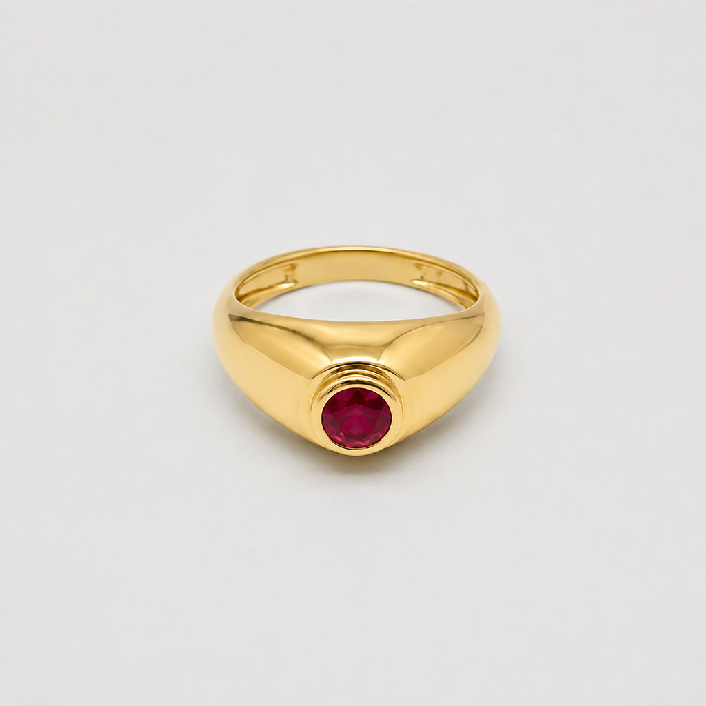 A polished yellow gold signet-style ring with a round deep red gemstone set in the center, shown on a clean light gray background.