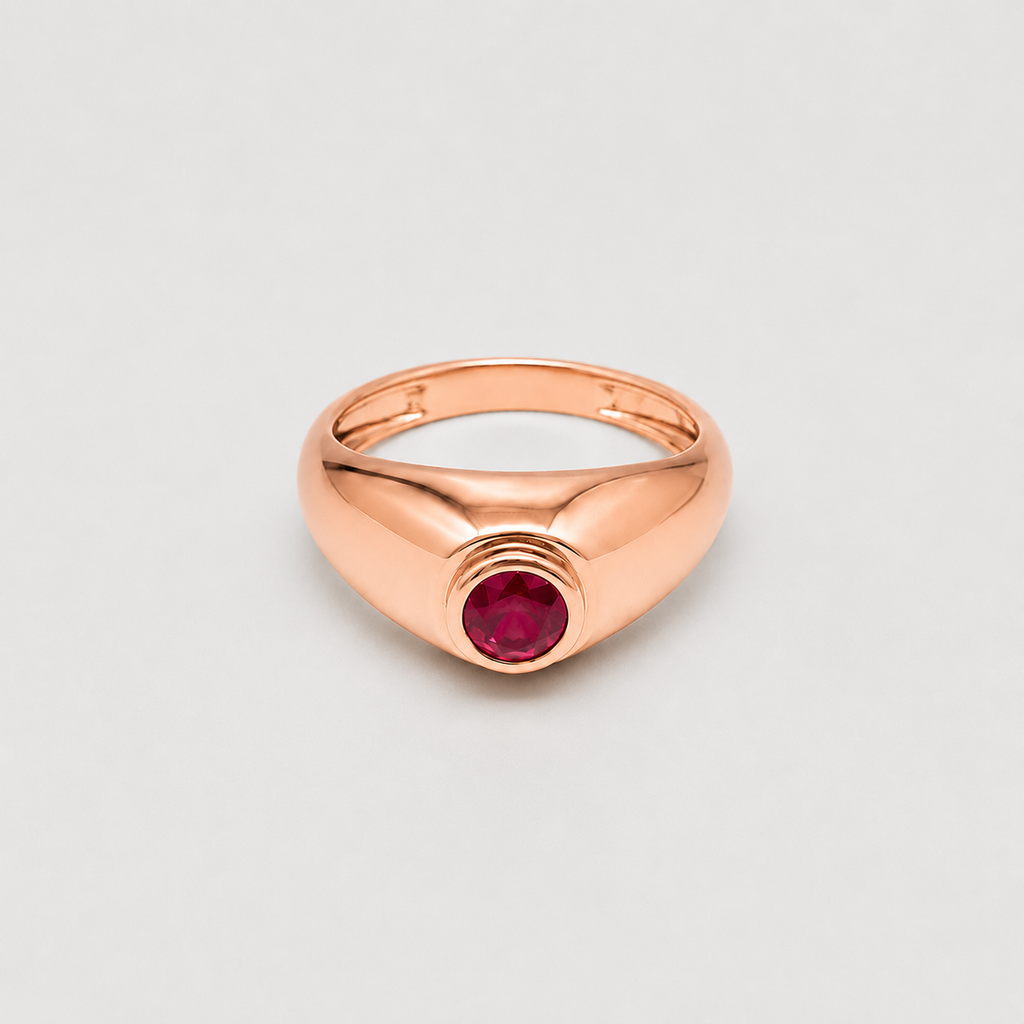 A rose gold signet-style ring with a smooth, polished band and a central round-cut dark red gemstone, shown on a light gray background.
