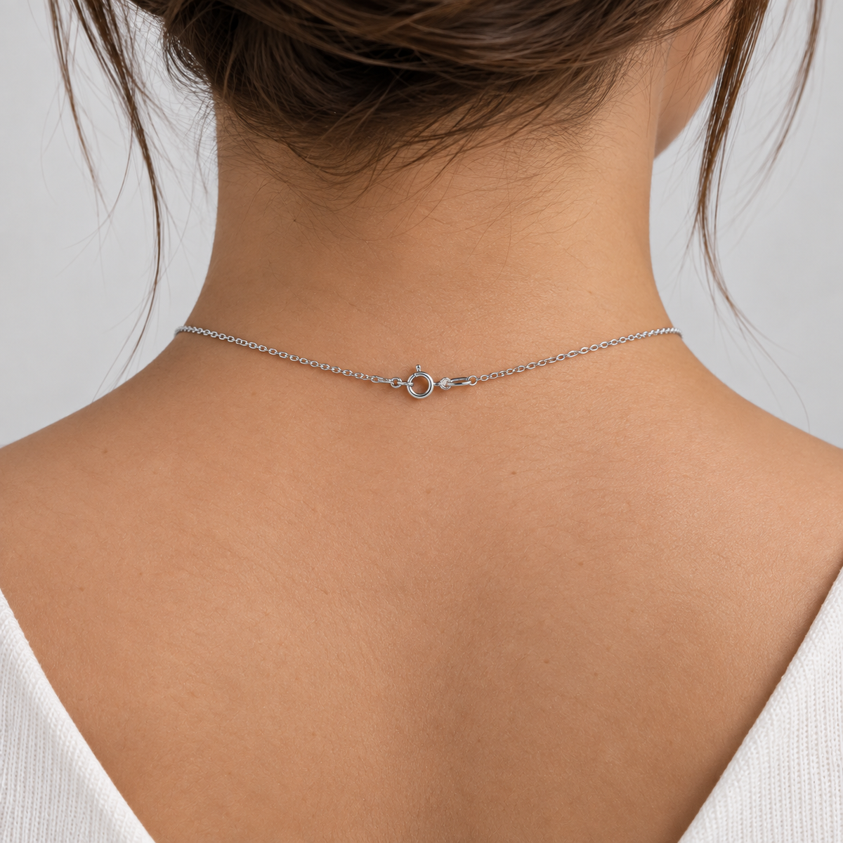 A close-up back view of a woman wearing a delicate silver chain necklace with a small circular charm at the center, shown against her neck and upper back.