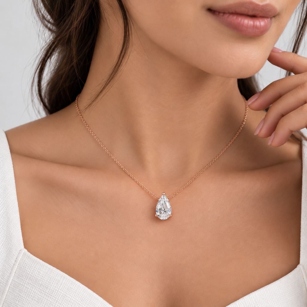 A close-up lifestyle shot of a model wearing a delicate rose gold chain necklace with a pear-cut clear gemstone pendant, styled with a white top against a soft light gray background.