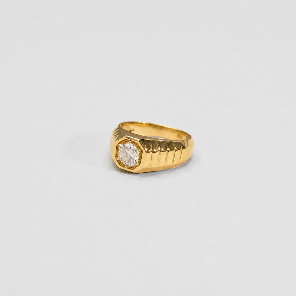 A yellow gold signet ring with a round-cut clear gemstone, set in a ribbed bezel and band, photographed on a light gray background.