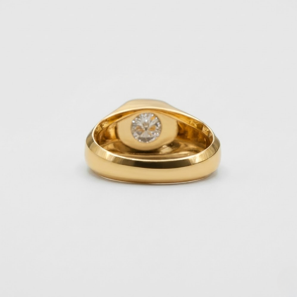A gold signet ring with a round-cut clear gemstone in the center, photographed on a clean light gray background.