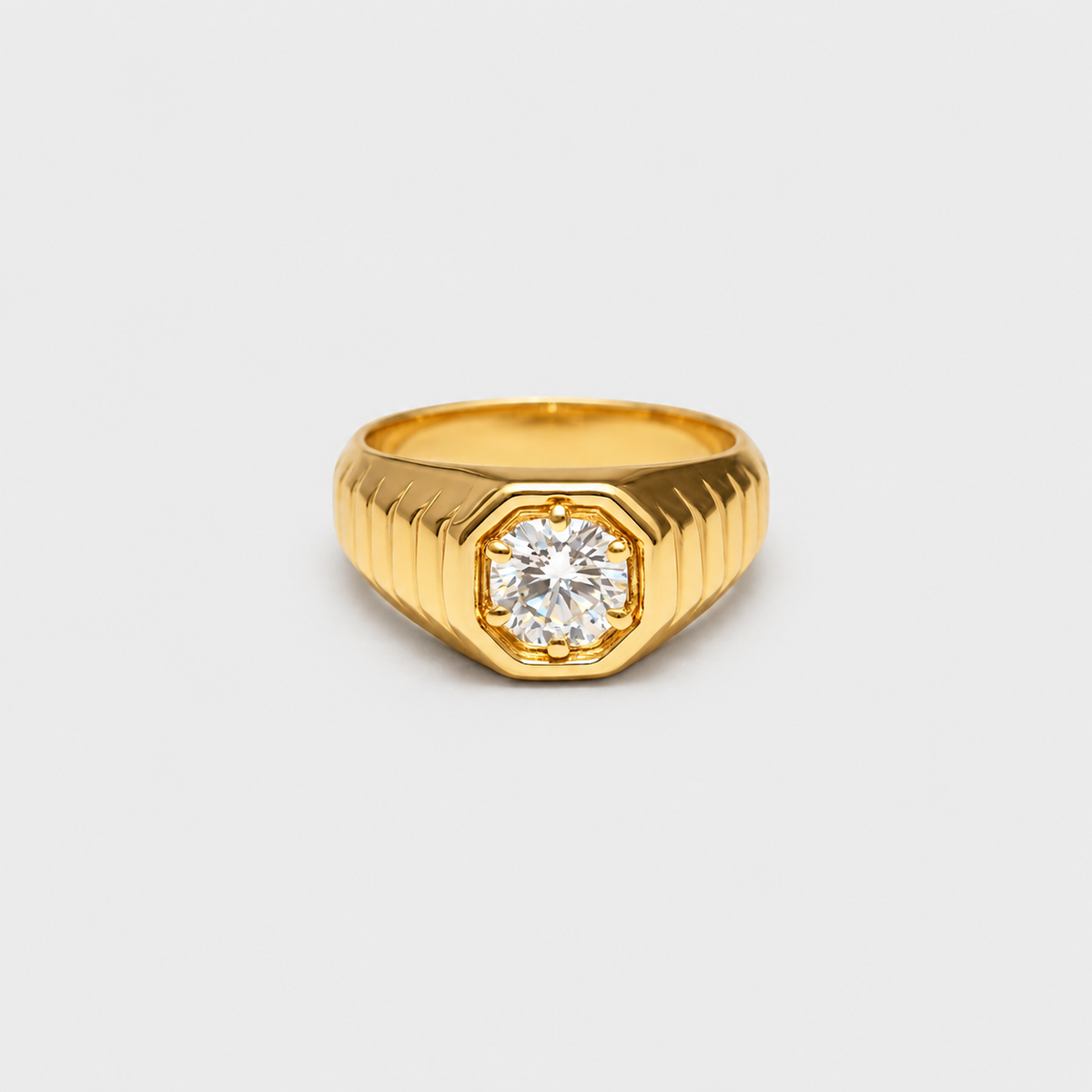 A gold signet-style ring with a faceted octagonal bezel holding a round clear gemstone, shown on a plain light gray background.