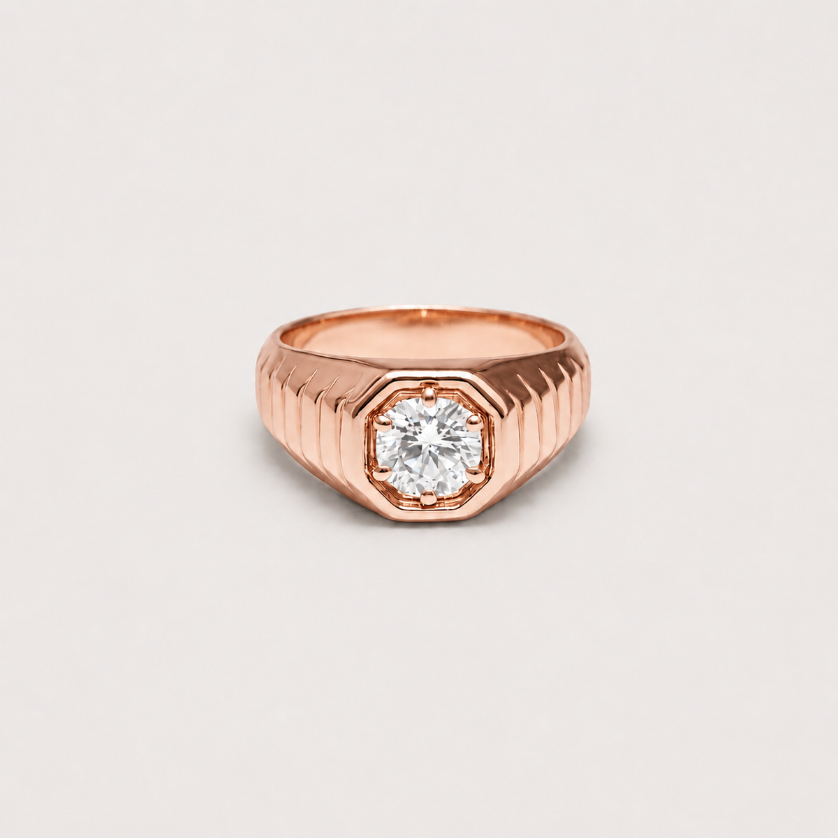 A rose gold signet-style ring with a faceted octagonal bezel and a clear round-cut center stone, shown on a light gray background.