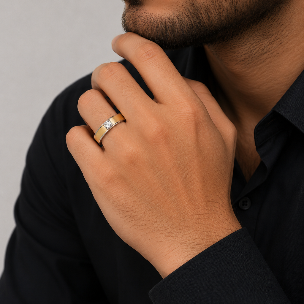 A close-up of a man's hand wearing a gold band ring with a small clear gemstone, set against a dark shirt and neutral gray background.