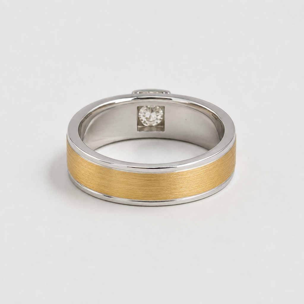 A two-tone metal ring with a brushed gold center band and polished silver-colored edges, featuring a small clear gemstone set in the center. The ring is photographed on a clean, light gray background.