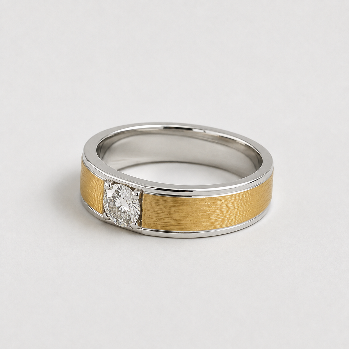 A two-tone ring band featuring a brushed gold center section, polished silver outer edges, and a single round-cut clear gemstone in a prong setting, photographed on a light gray background.