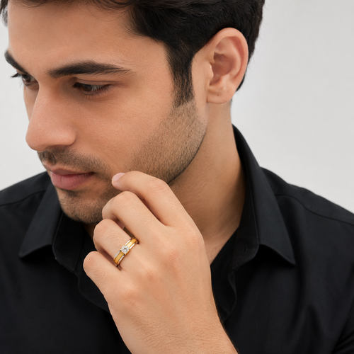 A close-up of a man wearing a black collared shirt and a gold ring with a small clear gemstone on his finger, set against a plain light gray background.