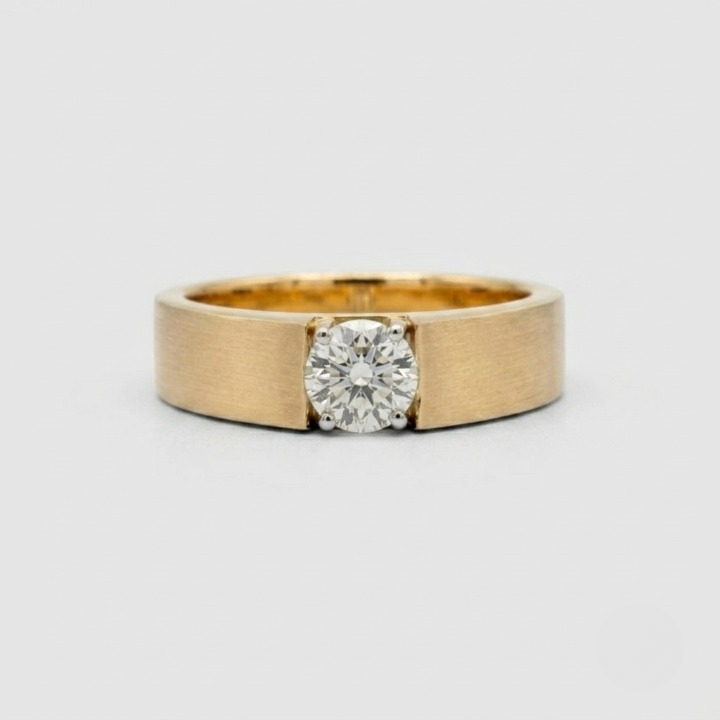 A yellow gold ring with a wide, brushed band and a round-cut clear gemstone in a four-prong setting, photographed on a light gray background.