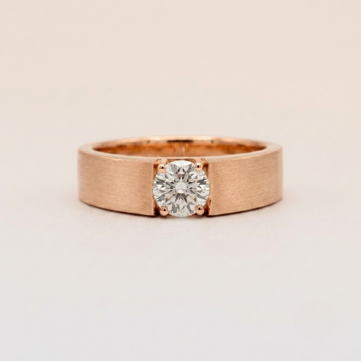 A minimalist rose gold band ring featuring a round-cut diamond set in a four-prong setting, photographed on a clean, light beige background.