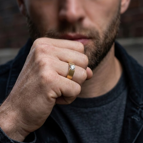 A close-up of a man wearing a gold band ring with a clear round gemstone on his ring finger, dressed in a dark denim jacket and gray t-shirt against a blurred brick wall background.