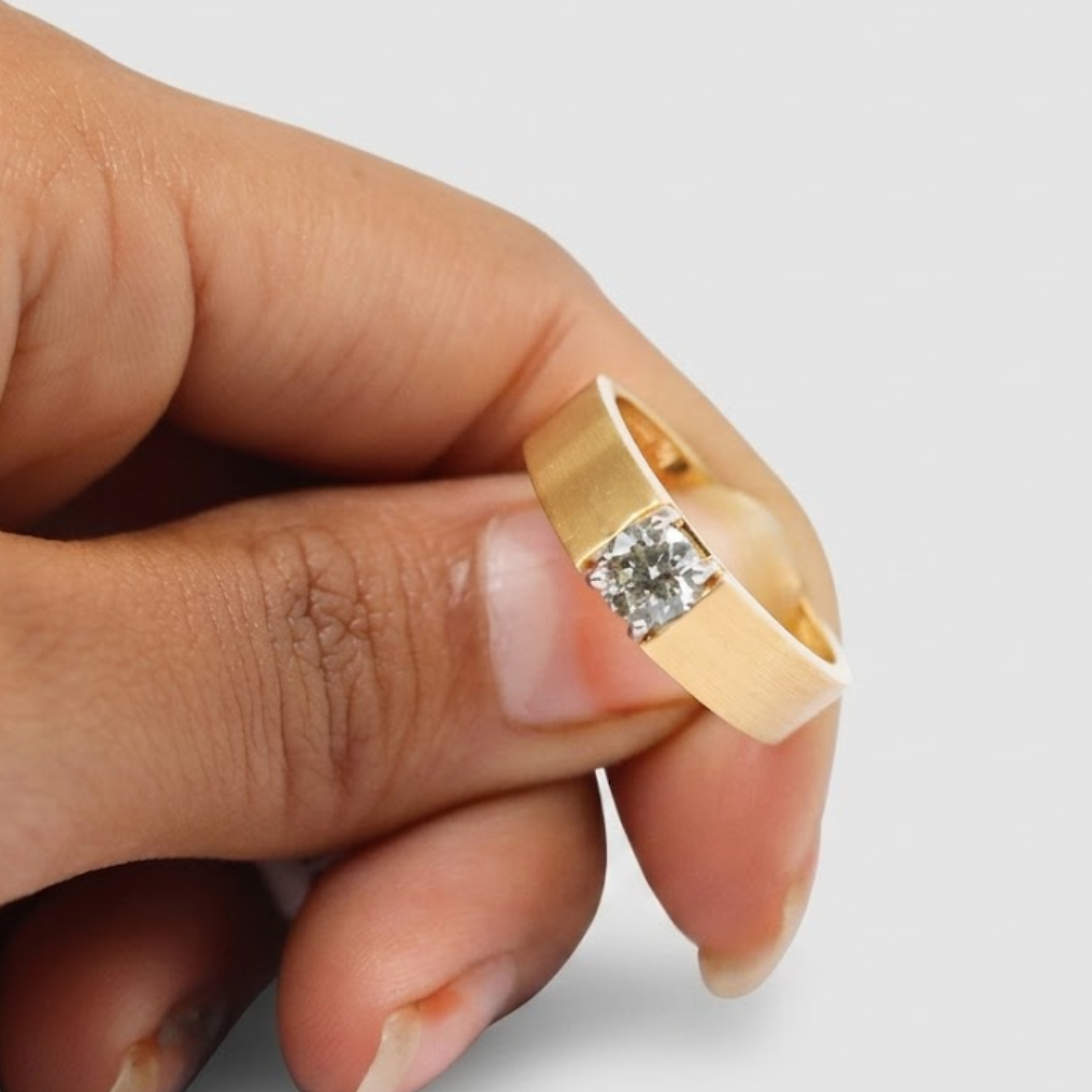 A gold ring with a single round clear gemstone held between fingers against a light gray background.