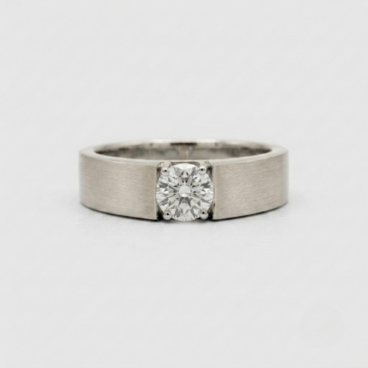 A minimalist solitaire ring featuring a round brilliant-cut diamond set in a brushed silver-toned metal band, photographed on a clean, light gray background.
