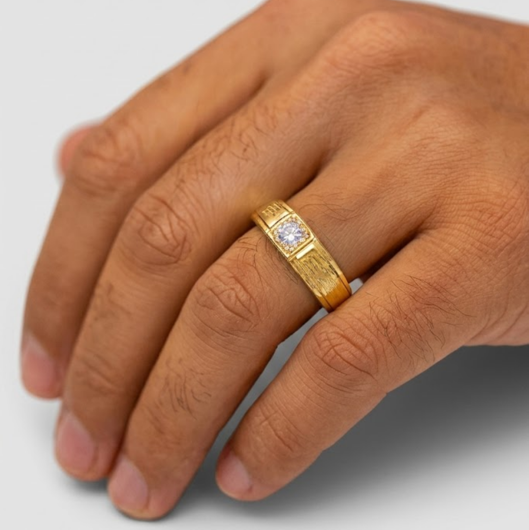 A gold band ring with a central clear gemstone, shown worn on a person's finger against a light gray background.