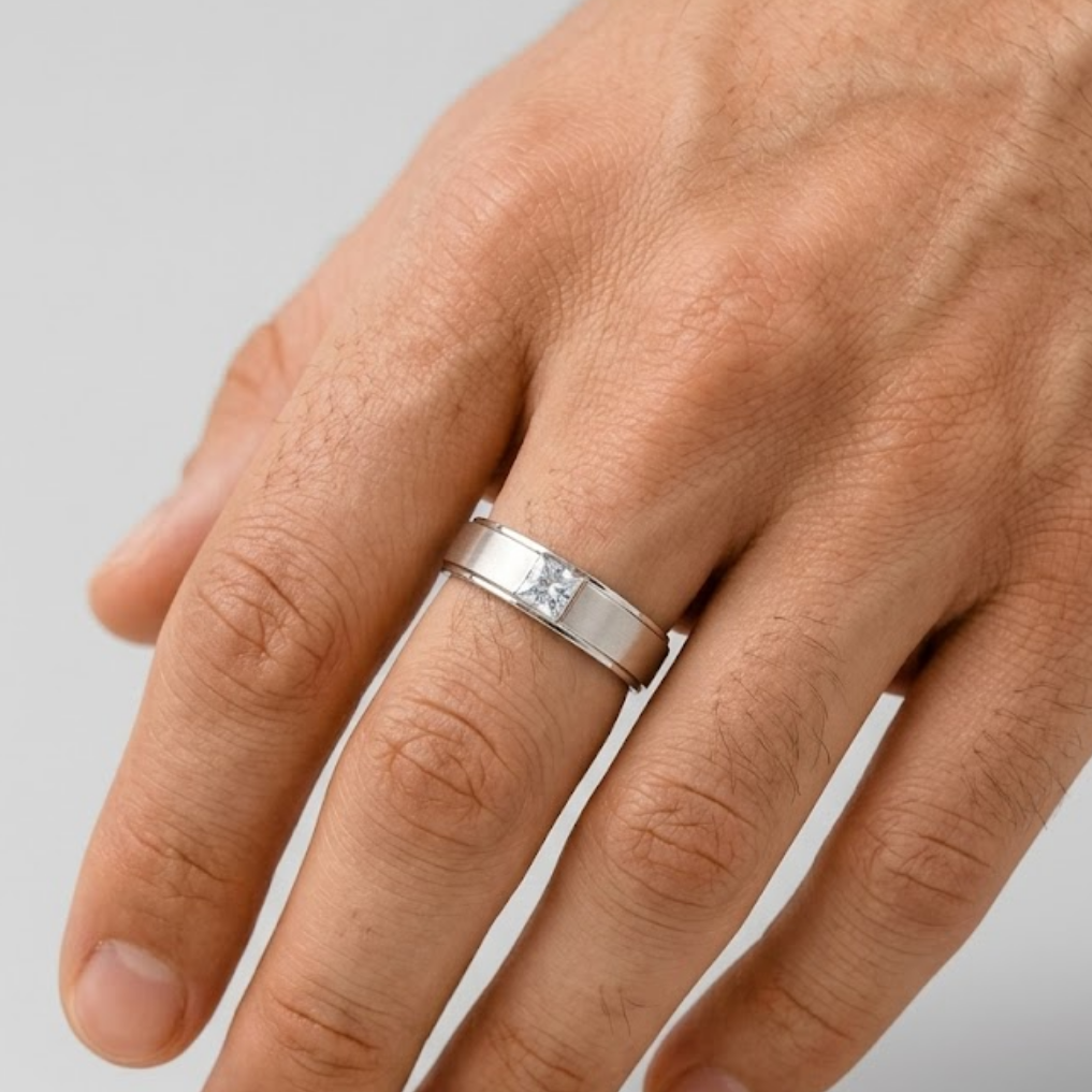 A silver band ring with a small clear gemstone set on a man's ring finger, shown against a neutral light gray background.