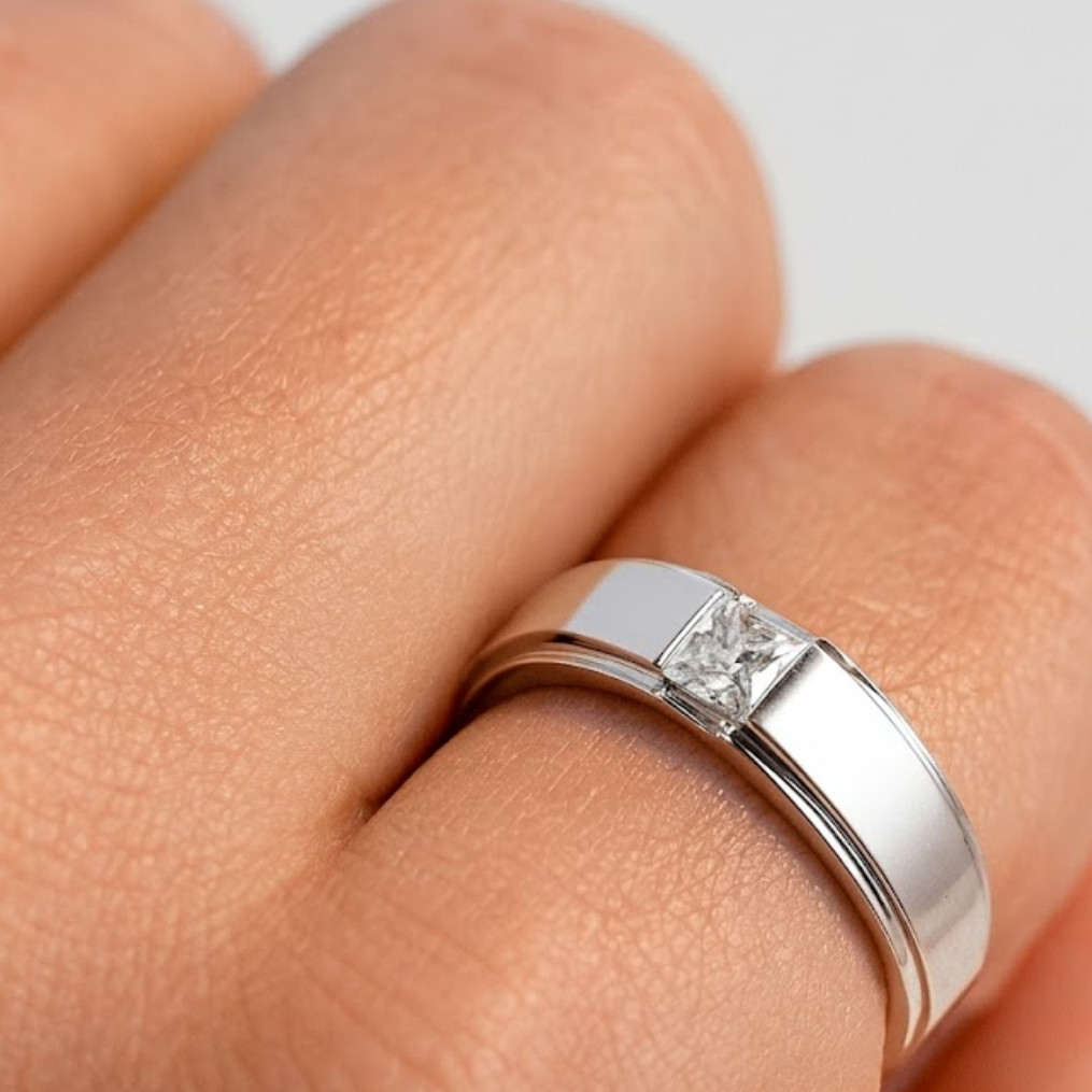 A silver band ring with a princess-cut diamond set in a four-prong setting, worn on a finger.