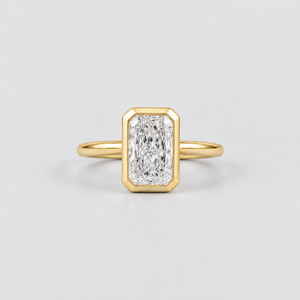 A yellow gold ring featuring an emerald-cut diamond in a beveled rectangular bezel setting, shown on a clean light gray background.