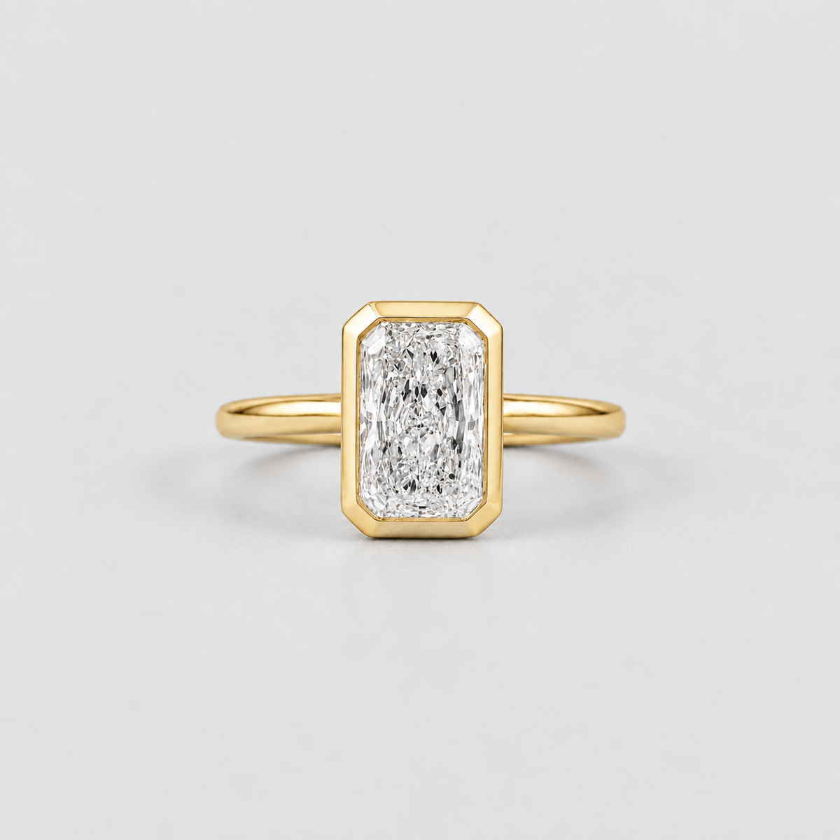A yellow gold ring featuring an emerald-cut diamond in a beveled rectangular bezel setting, shown on a clean light gray background.